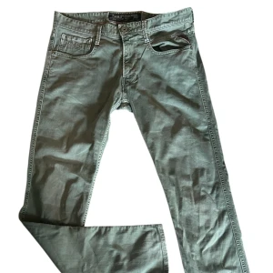 REPLAY Anbass 31 - Snygga grå jeansbyxor från Replay Denim med klassisk femficksdesign och diskreta röda detaljer vid fickan. Byxorna har raka ben och är tillverkade i ett mjukt bomullsmaterial med en skön passform. Perfekta för dig som gillar stilrena och bekväma jeans.