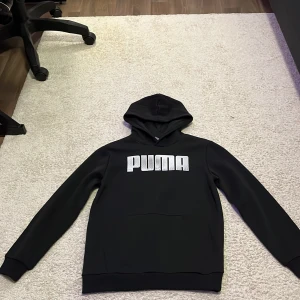 Svart hoodie från Puma med logga - Svart hoodie från Puma i storlek 164 med stor vit logga framtill. Klassisk modell med huva och magficka, tillverkad i mjuk bomullsblandning. Perfekt för dig som gillar sportig och clean stil. Tröjan är i mycket bra skick och den är inte använd någon gång. Priset kan diskuteras. 