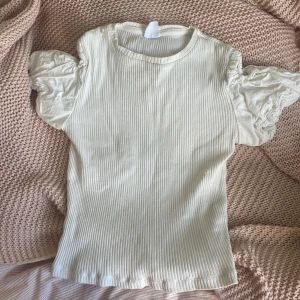 Vit ribbad topp med puffärmar - Supersöt vit topp med ribbad kropp och stora puffärmar i bomull. Ärmarna har öppen detalj vid axeln som ger en cool touch. Tight passform och rund halsringning. Perfekt för dig som gillar romantisk men trendig stil. Storlek 164cm 13-14år.