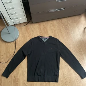 Svart v-ringad tröja Tommy Hilfiger S - Svart v-ringad tröja från Tommy Hilfiger i storlek S. Tillverkad i en lyxig mix av pima bomull och kashmir. Klassisk design med liten logga på bröstet och ribbade muddar. Perfekt för dig som gillar stilrena och bekväma plagg.