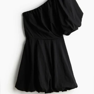 Svart oneshoulder kortklänning - Snygg svart kortklänning med oneshoulder-design och puffig ärm. Klänningen har en luftig passform och är perfekt för dig som vill sticka ut med en cool och trendig look. Tillverkad i ett lätt tyg som ger en skön känsla.