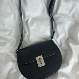 Svart axelväska med krokoprägling - Snygg svart axelväska med croco-mönstrad yta och silvrigt lås framtill. Väskan har rundad form, justerbar axelrem och ett rymligt huvudfack. Perfekt för att lyfta din outfit med en trendig detalj. Säljer pga den ej kommer till användning 