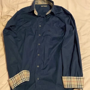 Blå Burberry skjorta - Snygg marinblå skjorta från Burberry i klassisk passform. Skjortan har långa ärmar, knappar framtill och ikoniska rutiga detaljer på insidan av kragen och ärmsluten. Tillverkad i bomull med lite stretch för extra komfort.