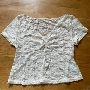 Vit spetsblus med v-ringning - Säljer en söt vit blus i spets med korta ärmar och v-ringning. Blusen har ett blommigt spetsmönster och är lätt transparent, perfekt för lager-på-lager. Den har en liten rynkdetalj framtill som ger extra stilpoäng.