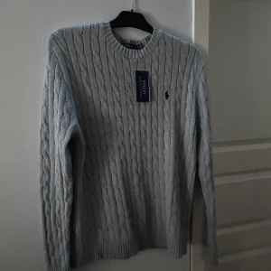 Grå stickad tröja från Ralph Lauren - Säljer en stilren grå stickad tröja från Ralph Lauren med kabelstickat mönster. Tröjan har långa ärmar och en klassisk rund halsring