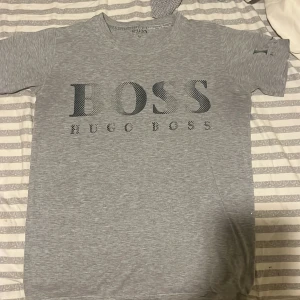 Grå t-shirt från Hugo Boss - Säljer en stilren grå t-shirt från Hugo Boss med deras logga tryckt på framsidan. Perfekt för en avslappnad look. T-shirten är kortärmad och gjord i ett mjukt material. Texten har suddats ut lite vid armen 