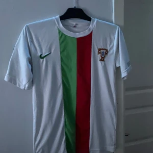 Vit fotbollströja med gröna och röda ränder - Säljer en vit fotbollströja från Nike med gröna och röda vertikala ränder. Tröjan har korta ärmar och ett FIFA World Cup 2010-märke på ärmen. Perfekt för fotbollsfans!