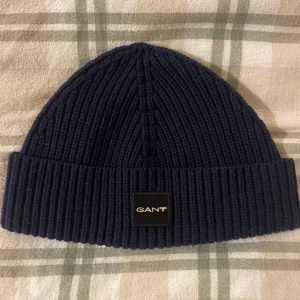 mössa från GANT - Snygg mörkblå ribbad mössa från GANT med en stilren logga framtill. Perfekt för att hålla värmen under kyliga dagar. Enkel och klassisk design som passar till det mesta.