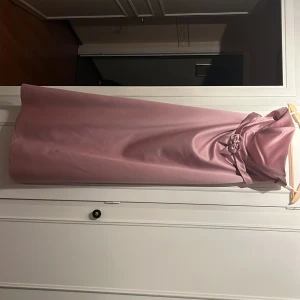 Ljusrosa axelbandslös klänning - Elegant ljusrosa axelbandslös klänning i satin med en dekorativ rosett i midjan. Perfekt för speciella tillfällen där du vill känna dig extra fin.