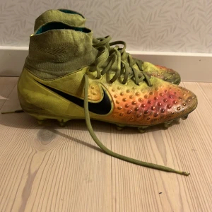 Färgglada fotbollsskor från Nike - Säljer ett par färgglada fotbollsskor från Nike med hög ankel och snörning. Skorna har en gradient av gult och orange med svarta detaljer och en Nike-logga på sidan. Perfekta för fotbollsplanen!