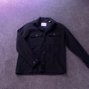 Svart overshirt med knappar - Säljer en stilren svart overshirt med knappar framtill och två bröstfickor. Perfekt för lager-på-lager-stil. Passar bra till både jeans och chinos.
