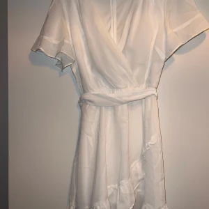 Studentklänning  - Säljer en elegant vit omlottklänning med korta ärmar och knytband i midjan. Klänningen har en v-ringad design och volangdetaljer vid fållen. Perfekt för studenten!!