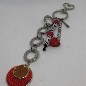 Nyckelring/väsksmycke med hjärtdetaljer och text - Charmig nyckelring/väsksmycke med flera detaljer, inklusive ett rött hjärta och en text som säger 'Love You'. Nyckelringen/väsksmycket har också flera silverfärgade ringar och en glittrig röd och orange cirkel. Perfekt som en personlig accessoar eller present.