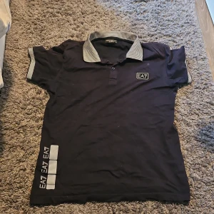 Svart pikétröja från Emporio Armani - Säljer en stilren svart pikétröja från Emporio Armani med randig krage och ärmslut. Tröjan har EA7-logga på bröstet och sidan. Perfekt för en casual look.