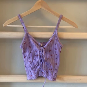 Lila ribbad linnetopp med blommönster - Söt lila linnetopp från Urban Outfitters med smala axelband, v-ringning och snörning framtill. Den har också med ett fint blommönster och är perfekt för varma sommardagar!🩷🎀💌