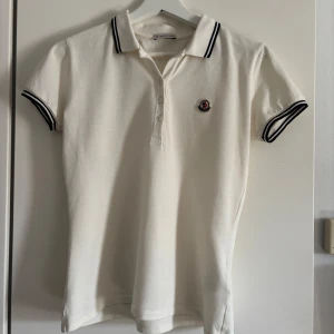 Vit pikétröja från Moncler - Säljer en stilren vit pikétröja från Moncler med mörkblå detaljer på krage och ärmslut. Tröjan har en klassisk pikédesign med knappar framtill och Monclers logotyp på bröstet.