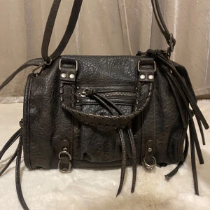 Svart axelväska med dragkedja - Snygg svart axelväska i skinn med flätade detaljer och dragkedja. Väskan har flera remmar och en praktisk ficka framtill. Perfekt för att bära dina dagliga nödvändigheter med stil.