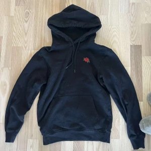 Svart hoodie med rosdetalj - Snygg svart hoodie med en liten broderad ros på bröstet. Den har en klassisk känguruficka och justerbar huva med dragsko. Perfekt för en avslappnad stil.