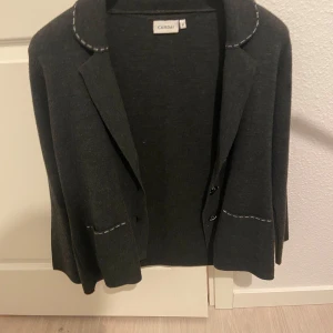 Mörkgrå kavaj från Canda - Snygg mörkgrå kavaj från Canda med dekorativa sömmar i vitt. Kavajen har knappar framtill och fickor med vita detaljer. Perfekt för en stilren look.