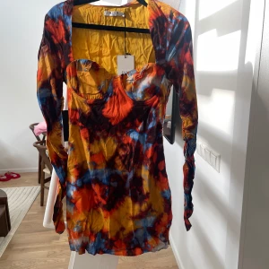 Färgglad klänning med långa ärmar - Säljer en färgglad klänning med långa ärmar och en unik batikmönster i orange, blått och gult. Klänningen har en tight passform och en vacker v-ringad design. Perfekt för att sticka ut med stil!