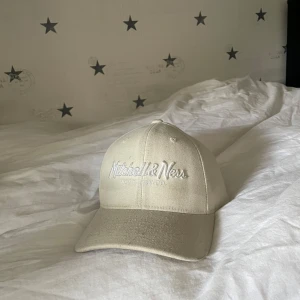 Beige keps från Mitchell & Ness - Snygg beige keps från Mitchell & Ness med broderad logga framtill. Kepsen har en klassisk design med justerbar rem baktill för perfekt passform. Perfekt för en avslappnad stil.