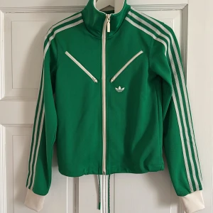Grön träningsjacka från Adidas - Snygg grön träningsjacka från Adidas med vita detaljer och dragkedja. Jackan har de klassiska tre ränderna längs ärmarna och två snedställda fickor med dragkedja framtill. Perfekt för en sportig look!