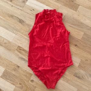 Röd ärmlös bodysuit från iEFiEL, stl M-L - Snygg röd ärmlös bodysuit från iEFiEL i ett glansigt material. Den har en hög krage med knappar i nacken och en öppen rygg. Perfekt för en stilren look. NY!!! Stl är en M-L