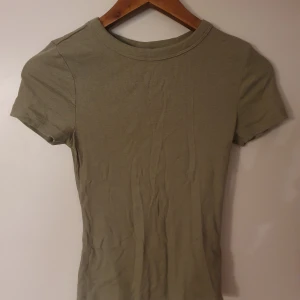 Olivgrön ribbad t-shirt - Säljer en olivgrön ribbad t-shirt med korta ärmar. Den har en tight passform och är perfekt för en avslappnad stil. T-shirten är enkel och stilren, vilket gör den lätt att matcha med olika outfits.
