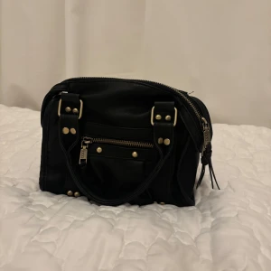 Svart handväska med dragkedja - Snygg svart handväska med dragkedja och dekorativa nitar. Väskan har dubbla handtag och en praktisk ytterficka med dragkedja. Perfekt för att hålla dina saker organiserade och lättillgängliga. Nästa helt oanvänd och i ny skick 