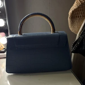 Blå handväska med krokodilmönster - Snygg blå handväska med krokodilmönster och ett elegant handtag i metall. Perfekt för att ge din outfit en stilfull touch. Väskan har en praktisk storlek för att bära dina nödvändigheter.
