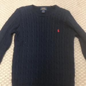 Mörkblå stickad tröja från Ralph Lauren - Säljer en klassisk mörkblå stickad tröja från Ralph Lauren med det ikoniska röda logotypbroderiet på bröstet. Tröjan har en kabelstickad design och långa ärmar, perfekt för kyligare dagar. Passar både till vardags och mer uppklädda tillfällen.