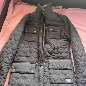 Svart quiltad jacka från Firetrap - Snygg svart quiltad jacka från Firetrap med dragkedja och knappar. Jackan har flera praktiska fickor och en hög krage för extra värme. Perfekt för kyligare dagar och ger en stilren look.