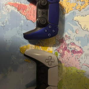 Blå och vit spelkontroll - Två spelkontroller, en i blått och en i vitt, ligger på en världskarta. De har en modern design med bekväma grepp och flera knappar för spelkontroll. Perfekt för gamingkvällar med vänner. Båda kontrollerna har stickdrift men det borde gå att lösa.