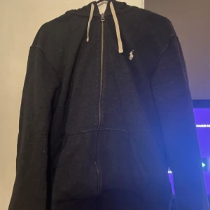 Svart hoodie från Polo Ralph Lauren - Säljer en svart hoodie från Polo Ralph Lauren med dragkedja och huva. Den har en liten broderad logga på bröstet och snören vid huvan. Perfekt för en avslappnad stil. Nypris 2400. 