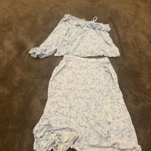 Blommigt set från Lindex - Säljer ett sött blommigt set från Lindex i ljusblått. Setet består av en offshoulder-topp med volangdetaljer och en matchande kjol. Perfekt för sommarens varma dagar!