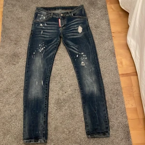 Blå jeans med slitningar - Säljer ett par blå jeans med coola slitningar och färgstänk. De har en klassisk femficksdesign och en röd detalj vid gylfen. Perfekta för en avslappnad stil.