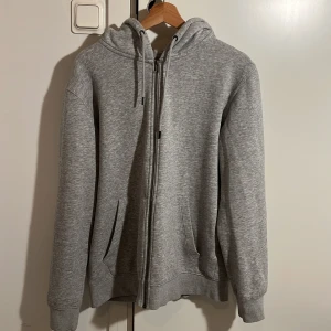 Grå hoodie med dragkedja - Säljer en stilren grå hoodie med dragkedja och justerbar huva. Tröjan har långa ärmar och praktiska fickor framtill. Perfekt för en avslappnad look.