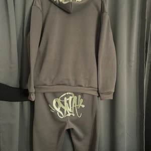 Grön Syna World tracksuit  - Säljer en grön Syna World tracksuit med tryck på huvan. Tröjan har en stor ficka framtill och snören vid huvan. Perfekt för en avslappnad stil. Endast testat 1 gång! 