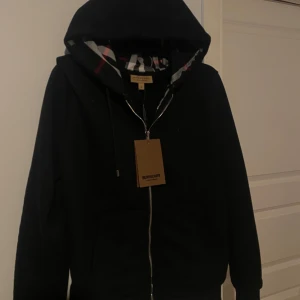 Svart hoodie från Burberry - Säljer en stilren svart hoodie från Burberry med dragkedja och klassiskt rutigt mönster i huvan. Tröjan har en broderad logga på bröstet och ribbade muddar vid ärmslut och nederkant. Perfekt för en casual look. Helt oanvänd, lappen sitter kvar på