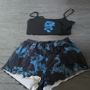 Svart pyjamas med blått drakmönster - Snygg svart pyjamas med ett coolt blått drakmönster. Toppen har smala axelband och byxorna har en liten rosett framtill. Perfekt för en sovstund.