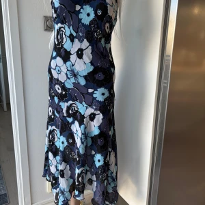 Blommig klänning figursydd - Säljer en vacker blommig klänning från KappAhl i olika nyanser av blått och svart. Klänningen har en v-ringad hals och är ärmlös, perfekt för sommarens festligheter. Den har en flödande passform med ett härligt mönster av stora blommor.