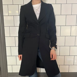 Svart kappa  - Elegant svart kappa med knappar framtill och klassisk krage. Perfekt för en stilren look. Långärmad design med en figursydd passform som ger en snygg siluett.