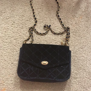 Mörka blå  axelväska från Holly & Whyte - Snygg svart axelväska i quiltad design från Holly & Whyte. Väskan har en elegant kedjerem och ett stilrent spänne i guld. Perfekt för att ge din outfit en lyxig touch.