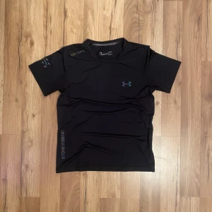 Tränings T-shirt från Under Armour - Säljer en svart kortärmad t-shirt från Under Armour med deras logga på bröstet. Tillverkad i ett lätt och andningsbart syntetmaterial, perfekt för träning. T-shirten har en rund hals och är designad för att hålla dig sval och bekväm under intensiva pass.