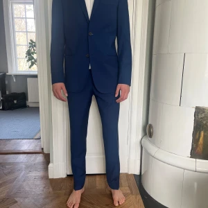 Blå kostym från J.Lindeberg - Snygg blå kostym från J.Lindeberg i slim fit. Kavajen har två knappar och är tillverkad i ett elegant material. Perfekt för formella tillfällen. Klädpåse ingår.