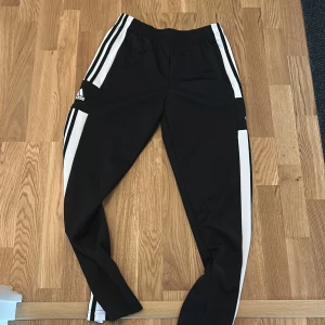 Svarta träningsbyxor från Adidas - Säljer ett par svarta träningsbyxor från Adidas med vita ränder längs sidorna. Byxorna har en elastisk midja och är perfekta för sport och träning. De är tillverkade i ett syntetmaterial som ger bra rörlighet.