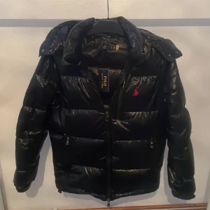 Polo Ralph Lauren - Snygg svart pufferjacka från Polo Ralph Lauren med en röd logotyp på bröstet. Jackan har en dragkedja framtill och en avtagbar huva. Perfekt för kyliga dagar.