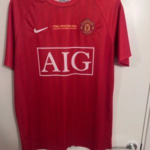 08 Manchester United fotbollströja - Säljer en röd fotbollströja från Nike med Manchester Uniteds emblem och AIG-logga på framsidan. Texten 'FINAL MOSCOW 2008' är tryckt ovanför loggan. På baksidan står 'RONALDO' och nummer 7. Tröjan har korta ärmar och är perfekt för fotbollsfans.