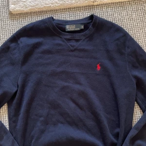 Mörkblå tröja från Polo Ralph Lauren - Säljer en stilren mörkblå tröja från Polo Ralph Lauren med det klassiska röda logotypbroderiet på bröstet. Tröjan har långa ärmar och en rund halsringning. Perfekt för en avslappnad och snygg look.