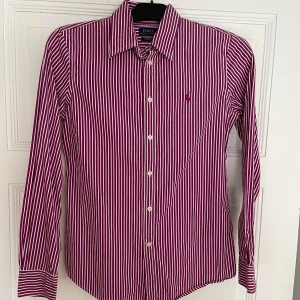Randig skjorta från Ralph Lauren - Snygg randig skjorta från Ralph Lauren i vinrött och vitt. Skjortan har långa ärmar och knappar framtill. Den är broderad med den klassiska logotypen på bröstet. Perfekt för en stilren look!
